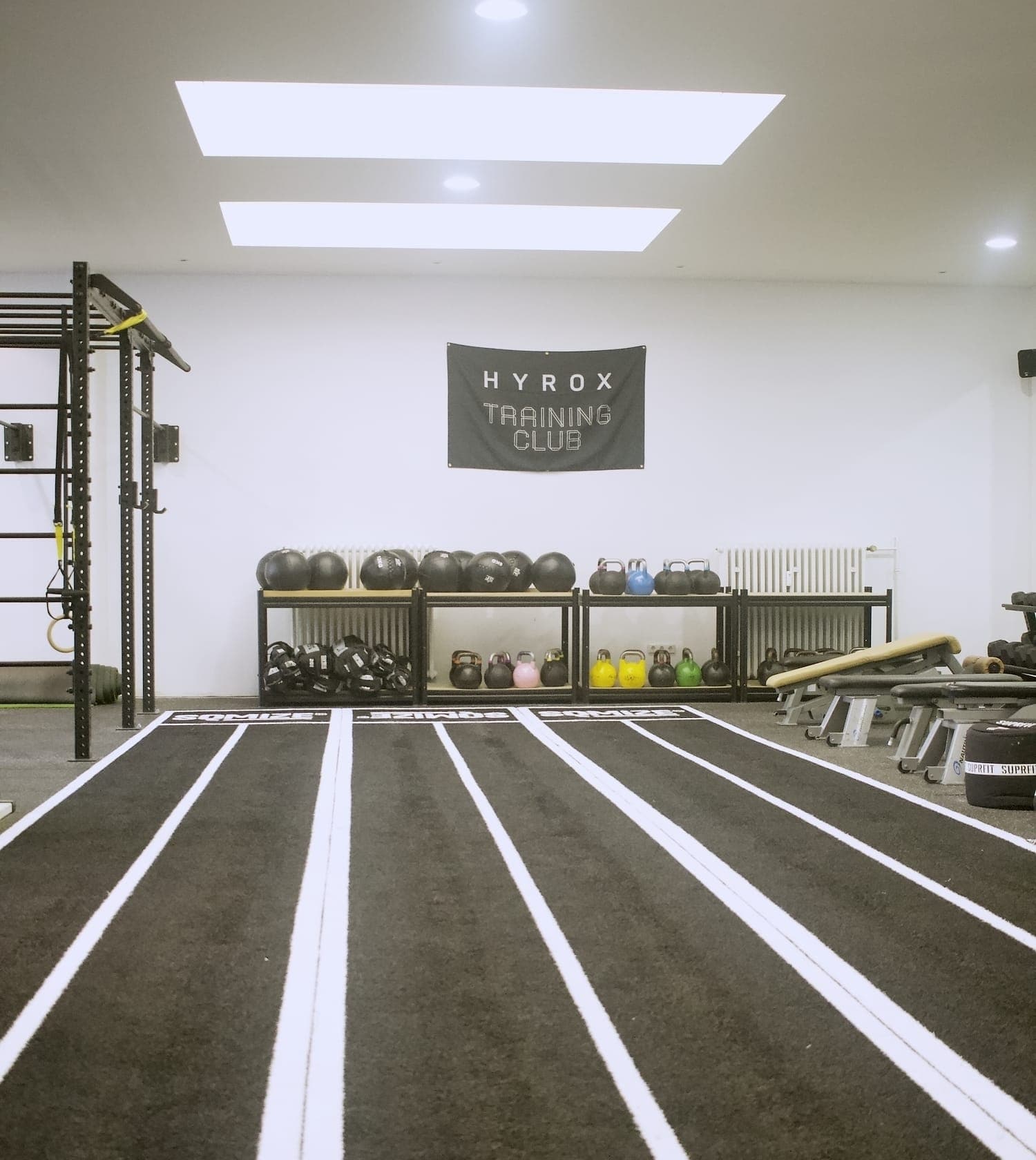 Drei markierte Bahnen laufen auf die Wand mit dem HYROX-Banner zu, davor Wall Balls und Kettlebells im Regal